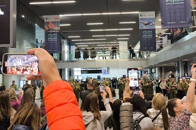 muzycy orkiestry reprezentacyjnej straży granicznej podczas kręcenia flash moba na krakowskim lotnisku. obserwują ich zebrani podróżni. używają telefonów aby zrejestrować występ orkiestry reprezentacyjnej straży granicznej.