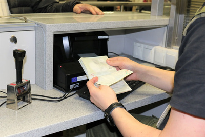 wnętrze kabiny kontrolerskiej , funkcjonariusz sg sprawdza paszport podróżnego. w tle komputer i urządzenia do weryfikacji autentyczności dokumentów