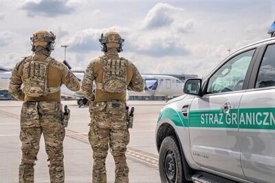 Dwaj funkcjonariusze ZIS PSG Kraków - Balice ustawieni tyłem do zdjęcia w kamizelkach taktycznych z napisem straż graniczna, a twarzą zwróceni w stronę portu lotniczego.