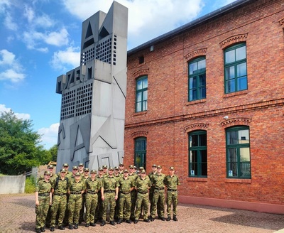 nowo przyjęci funkcjonariusze straży granicznej pozują do wspólnego zdjęcia przed budynkiem muzeum armii krajowej w Krakowie. budynek jest zbudowany z czerwonej cegły. za nimi stoi pomnik z napisem AK poniżej jest napis Muzeum