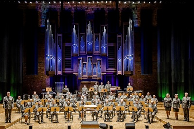 orkiestra reprezentacyjna straży granicznej pozująca do pamiątkowego zdjęcia w filharmonii. po prawej stronie orkiestry stoją soliści orkiestry (dwie funkcjonariuszki oraz funkcjonariusz), natomiast po lewej stronie orkiestry stoi kapelmistrz orkiestry.
