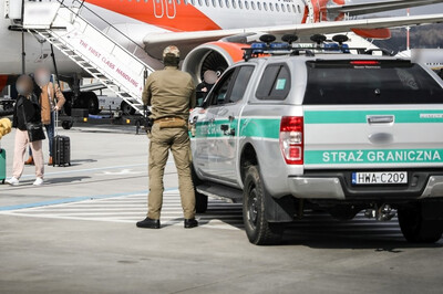 pojazd służbowy straży granicznej z napisem straż graniczna na tylnej klapie i bocznych drzwiach stoi na płycie lotniska. obok pojazdu stoi funkcjonariusz straży granicznej obserwujący pasażerów, którzy wysiadają z samolotu.