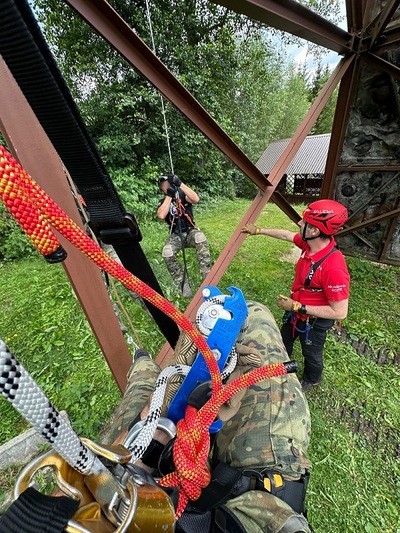 Jeden funkcjonariusz w kasku i pełnym oprzyżądowaniu wisi na linie, spuszczonej z wieży wysokościowej. Instruktażu udziela mu ratownik GOPR.