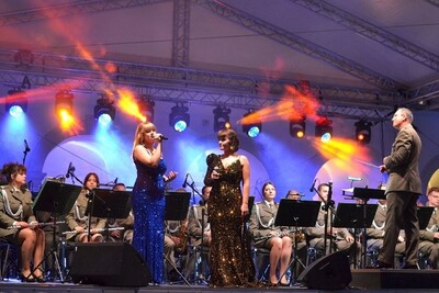 Na scenie dwie wokalistki w wieczorowych sukniach śpiewają za nimi orkiestra gra a obok dyrygent kieruje orkiestrą