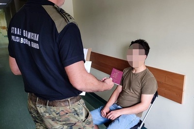 Zatrzymany cudzoziemiec siedzi na krześle. Przed nim stoi funkcjonariusz straży granicznej, który trzyma w rękach paszport i dokumenty z przesłuchania cudzoziemca.