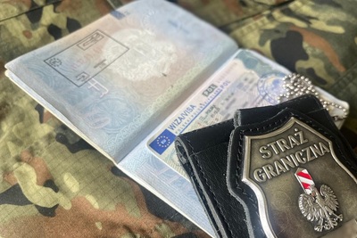 Na materiale w kolorze moro leży otwarty paszport z podrobioną wizą i stemplem kontroli granicznej oraz odznaka metalowa funkcjonariusza SG.
