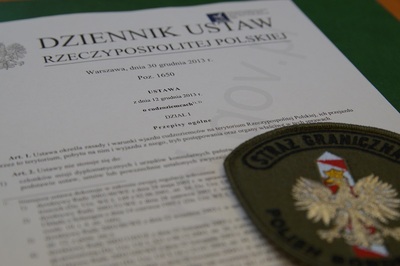 emblemat straży granicznej koloru khaki z napisem straż graniczna oraz widocznym orłem biały, który jest usytuowany na białoczerwonym słupku granicznym. emblemat znajduje się na papierowej wersji ustawy o cudzoziemcach.