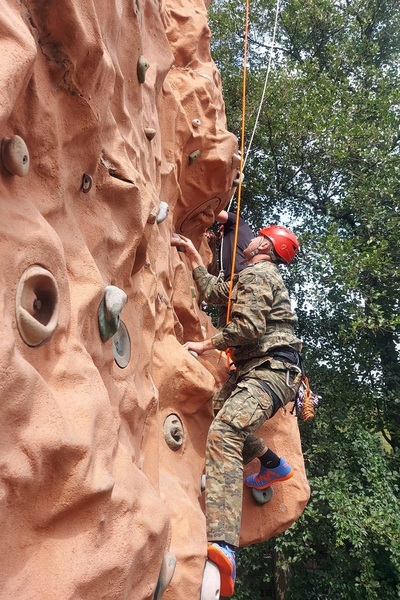 Funkcjonariusz SG wspina się na ćwiczeniowej ściance wspinaczkowej w kolorze brązowym. Ma na głowie kask w kolorze czerwonym. Wyposażony jest również w sprzęt wspinaczkowy.