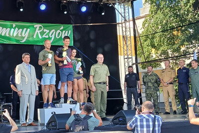 funkcjonariusze straży granicznej stojący na podium podczas ceremonii wręczenia medali po zakończonych zawodach. w tle stoją dowódcy poszczególnych jednostek wojska, policji straży pożarnej. uroczystość odbywa się na scenie w plenerze.