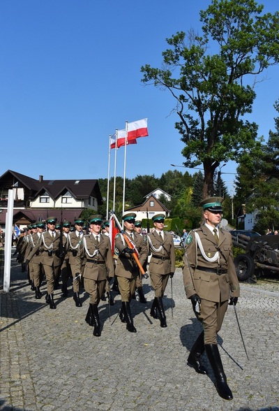 węgierska górka. kompania reprezentacyjna sg kroczy krokiem defiladowym w zwartym szyku, przed nimi idzie dowódca z szablą w dłoni. w tle powiewają trzy biało czerwone flagi na masztach