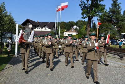 krokiem defiladowym idzie grająca na instrumentach orkiestra reprezentacyjna sg prowadzona przez tamburmajora z buławą w rękach. w tle powiewają biało czerwone flagi na trzech masztach