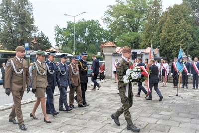 żołnierz trzymający na rękach wieniec złożony z białoczerwonych kwiatów niosący go pod pomnik podczas uroczystości związanych z wybuchem II wojny światowej. za żołnierzem idą przedstawiciele służb mundurowych Straży granicznej, policji, wojska, straży pożarnej, służby więziennej.