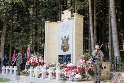 na zdjęciu widoczny wielki pomnik w kolorze białym poświęconym poległym żołnierzom. przed pomnikiem wystawiona jest warta honorowa złożona z dwóch żołnierze. przed pomnikiem złożone są kwiaty i wieńce.