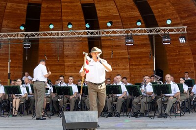 solista orkiestry reprezentacyjnej straży granicznej występujący podczas koncertu. za nim siedzą muzycy orkiestry reprezentacyjnej straży granicznej grający podczas koncertu w amfiteatrze. funkcjonariusze ubrani są w białe służbowe koszule z pagonami oraz spodnie koloru khaki. dyryguje nimi ich naczelnik