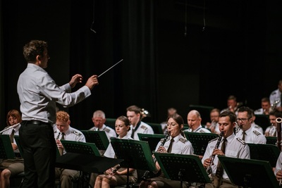 dyrygent adept podczas egzaminu z dyrygowania dyryguje orkiestrą reprezentacyjną straży granicznej. wydarzenie odbywa się w sali koncertowej.