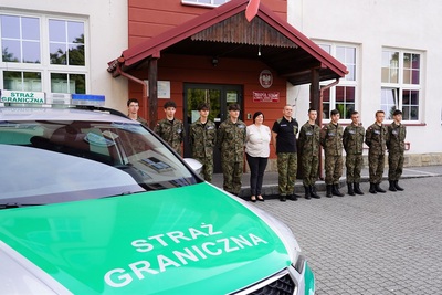 uczniowie klasy mundurowej o profilu straż graniczna pozujący do wspólnego zdjęcia wraz z dyrektorka szkoły oraz nauczycielem który jest funkcjonariuszem straży granicznej. na pierwszym planie pojazd służbowy z napisem straż graniczna.