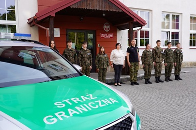 uczniowie klasy mundurowej o profilu straż graniczna pozujący do wspólnego zdjęcia wraz z dyrektorka szkoły oraz nauczycielem który jest funkcjonariuszem straży granicznej. na pierwszym planie pojazd służbowy z napisem straż graniczna.