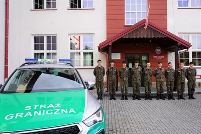 uczniowie klasy mundurowej o profilu straż graniczna pozujący do wspólnego zdjęcia wraz z dyrektorka szkoły oraz nauczycielem który jest funkcjonariuszem straży granicznej. na pierwszym planie pojazd służbowy z napisem straż graniczna.