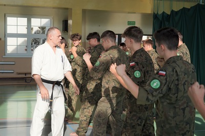 uczniowie klasy mundurowej o profilu straż graniczna wykonują ćwiczenia z samoobrony. prowadzi je funkcjonariusz straży granicznej ubrany w kimono karate w kolorze białym z czarnym pasem.