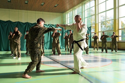 uczeń klasy mundurowej o profilu straż graniczna wykonuje ćwiczenie z samoobrony. prowadzi je funkcjonariusz straży granicznej ubrany w kimono karate w kolorze białym z czarnym pasem. reszta uczniów na drugim planie przypatruje się i również wykonuje ćwiczenie.