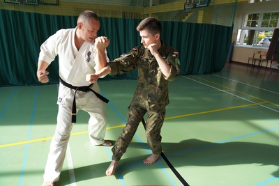 uczeń klasy mundurowej o profilu straż graniczna wykonuje ćwiczenie z samoobrony. prowadzi je funkcjonariusz straży granicznej ubrany w kimono karate w kolorze białym z czarnym pasem. reszta uczniów na drugim planie przypatruje się i również wykonuje ćwiczenie.