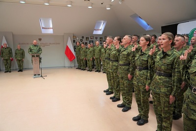 nowoprzyjęci funkcjonariusze straży granicznej składają ślubowanie. Mają podniesione prawe ręce i wystawione dwa złączone palce do ślubowania. Rotę ślubowania czyta komendant Karpackiego oddziału straży granicznej. W tle stoi kadra kierownicza oddziału. Widoczna jest również białoczerwona flaga.