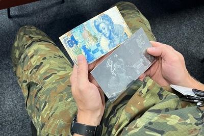 Funkcjonariusz trzyma w rękach wzór banknotu oraz matrycę do wykonania tego banknotu.