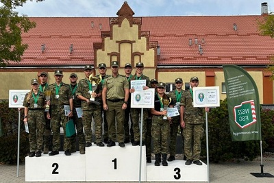 Na podium stoją w towarzystwie komendanta warmińsko-mazurskiego oddziału sg trzy zwycięskie drużyny. w rękach trzymają puchary i dyplomy oraz tabliczki z oznczeniem nazwy oddziału z którego pochodzą