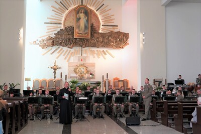 ksiądz proboszcz przemawia do zebranej publiczności podczas koncertu. obok stoi kapelmistrz orkiestry. za nimi siedzą funkcjonariusze straży granicznej, którzy są muzykami i służą w orkiestrze reprezentacyjnej straży granicznej. na drugim planie widoczny jest ołtarz a na ścianie widoczny obraz Jezu Ufam Tobie.