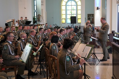 orkiestra reprezentacyjna straży granicznej podczas koncertu charytatywnego. przed orkiestrą dyryguje jej kapelmistrz. wszyscy ubrani są w mundur wyjściowy w kolorze khaki. koncert odbywa się w kościele.