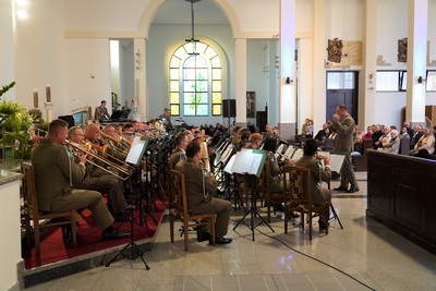 orkiestra reprezentacyjna straży granicznej podczas koncertu charytatywnego. przed orkiestrą dyryguje jej kapelmistrz. wszyscy ubrani są w mundur wyjściowy w kolorze khaki. koncert odbywa się w kościele. widoczna również jest publiczność siedząca w ławach kościoła.