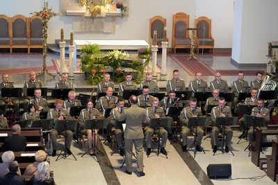 orkiestra reprezentacyjna straży granicznej podczas koncertu charytatywnego. przed orkiestrą dyryguje jej kapelmistrz. wszyscy ubrani są w mundur wyjściowy w kolorze khaki. koncert odbywa się w kościele. za orkiestrą widoczny jest ołtarz. widoczna również jest publiczność siedząca w ławach kościoła.