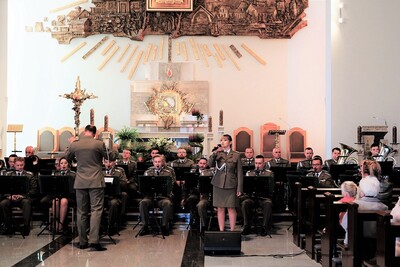 orkiestra reprezentacyjna straży granicznej podczas koncertu charytatywnego. przed orkiestrą dyryguje jej kapelmistrz. przed orkiestra a obok kapelmistrza stoi  wokalistka, która wykonuje utwór wokalnie. wszyscy ubrani są w mundur wyjściowy w kolorze khaki. koncert odbywa się w kościele. za orkiestrą widoczny jest ołtarz. widoczna również jest publiczność siedząca w ławach kościoła.