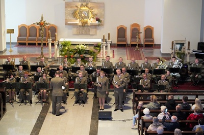 orkiestra reprezentacyjna straży granicznej podczas koncertu charytatywnego. przed orkiestrą dyryguje jej kapelmistrz. przed orkiestra a obok kapelmistrza stoi  wokalistka wraz z wokalistą, którzy wykonują utwór wokalnie. wszyscy ubrani są w mundur wyjściowy w kolorze khaki. koncert odbywa się w kościele. za orkiestrą widoczny jest ołtarz. widoczna również jest publiczność siedząca w ławach kościoła.