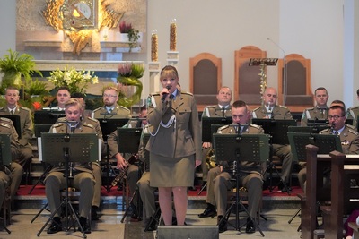 orkiestra reprezentacyjna straży granicznej podczas koncertu charytatywnego. przed orkiestrą  stoi  wokalistka, która wykonuje utwór wokalnie. wszyscy ubrani są w mundur wyjściowy w kolorze khaki. koncert odbywa się w kościele. za orkiestrą widoczny jest ołtarz. widoczna również jest publiczność siedząca w ławach kościoła.