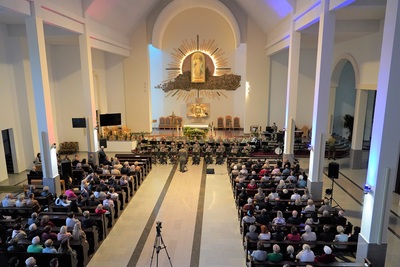 orkiestra reprezentacyjna straży granicznej podczas koncertu charytatywnego. przed orkiestrą dyryguje jej kapelmistrz. wszyscy ubrani są w mundur wyjściowy w kolorze khaki. koncert odbywa się w kościele. za orkiestrą widoczny jest ołtarz oraz na ścianie widoczny jest obraz Jezu Ufam Tobie. widoczna również jest publiczność siedząca w ławach kościoła.