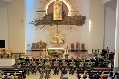 orkiestra reprezentacyjna straży granicznej podczas koncertu charytatywnego. przed orkiestrą dyryguje jej kapelmistrz. wszyscy ubrani są w mundur wyjściowy w kolorze khaki. koncert odbywa się w kościele. za orkiestrą widoczny jest ołtarz oraz na ścianie widoczny jest obraz Jezu Ufam Tobie. widoczna również jest publiczność siedząca w ławach kościoła.