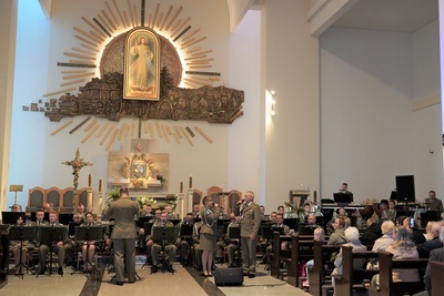 orkiestra reprezentacyjna straży granicznej podczas koncertu charytatywnego. przed orkiestrą dyryguje jej kapelmistrz. przed orkiestrą a obok kapelmistrza stoi  wokalistka oraz wokalista, którzy wykonują utwór wokalnie. wszyscy ubrani są w mundur wyjściowy w kolorze khaki. koncert odbywa się w kościele. za orkiestrą widoczny jest ołtarz oraz na ścianie widoczny jest obraz Jezu Ufam Tobie. widoczna również jest publiczność siedząca w ławach kościoła.