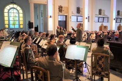 orkiestra reprezentacyjna straży granicznej podczas koncertu charytatywnego. przed orkiestrą dyryguje jej kapelmistrz. przed orkiestrą a obok kapelmistrza stoi  wokalistka oraz wokalista, którzy wykonują utwór wokalnie. wszyscy ubrani są w mundur wyjściowy w kolorze khaki. koncert odbywa się w kościele. widoczna również jest publiczność siedząca w ławach kościoła.