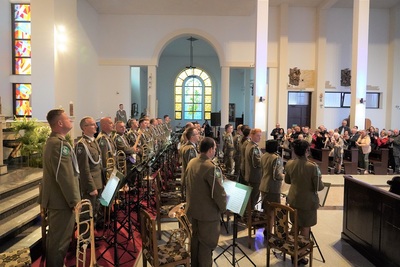 orkiestra reprezentacyjna straży granicznej podczas koncertu charytatywnego.  wszyscy ubrani są w mundur wyjściowy w kolorze khaki. koncert odbywa się w kościele.  widoczna również jest publiczność siedząca w ławach kościoła.