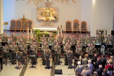 orkiestra reprezentacyjna straży granicznej podczas koncertu charytatywnego. przed orkiestrą kłaniają się do zebranej publiczności kapelmistrz oraz wokaliści orkiestry, wszyscy ubrani są w mundur wyjściowy w kolorze khaki. koncert odbywa się w kościele. za orkiestrą widoczny jest ołtarz. widoczna również jest publiczność.