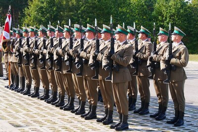 kompania reprezentacyjna straży granicznej. funkcjonariusze stoją dwuszeregu na baczność. ubrani są w mundury wyjściowe w kolorze khaki. w rękach trzymają karabiny reprezentacyjne.  w oddali widoczny jest sztandar.