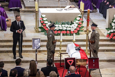 na pierwszym planie trumna na której położona jest flaga białoczerwona. obok trumny stoją funkcjonariusze straży granicznej w postawie na baczność. widoczne są kwiaty białoczerwone oraz świece. po lewej strony z ambony przemawia do zebranych prezydent Polski. uroczystość odbywa się w kościele.