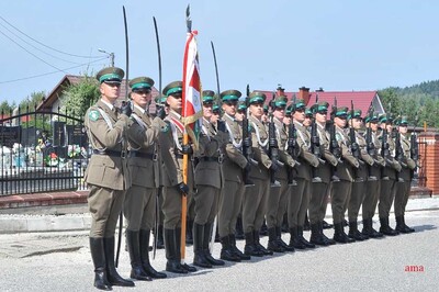 kompania reprezentacyjna straży granicznej stojąca w szyku na baczność. po lewej stronie stoi dowódca kompanii następnie poczet sztandarowy ze sztandarem oraz funkcjonariusze straży granicznej którzy tworzą kompanie . trzymają karabiny reprezentacyjne natomiast dowódca i poczet sztandarowy w dłoniach mają szable.