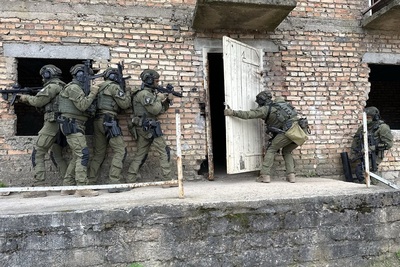 funkcjonariusze straży granicznej wykonujący ćwiczenia przy budynku na poligonie wojskowym. na zdjęciu widoczni funkcjonariusze podchodzący pod budynek w zwartej grupie. otwierają białe drzwi budynku.  są wyposażeni w broń i umundurowani na bojowo.