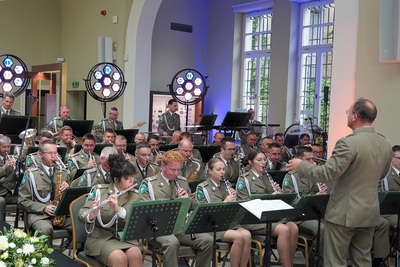 na pierwszym planie widoczny dyrygent orkiestry reprezentacyjnej straży granicznej, który dyryguje orkiestrą. przed nim siedzą muzycy orkiestry. koncert odbywa się w pomieszczeniu.