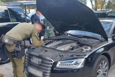 funkcjonariusz straży granicznej sprawdzający pojazd na materiały wybuchowe i niebezpieczne. sprawdza pojazd za pomocą latarki aby dostrzec trudnodostępne miejsca pod maska pojazdu. widoczny jest silnik pojazdu oraz podniesiona maska pojazdu. całość odbywa się na zewnątrz.