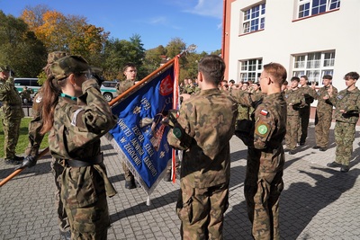 czterech uczniów klasy mundurowej ślubuję na sztandar swojej szkoły. za nimi reszta uczniów ślubuje na sztandar szkoły. całość odbywa się na zewnątrz. w tle budynek szkoły.