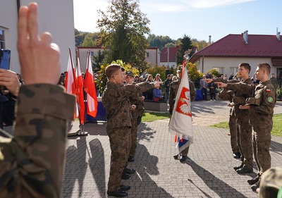 czterech uczniów klasy mundurowej ślubuję na sztandar swojej szkoły. całość odbywa się na zewnątrz. w tle widoczni ludzie zebrani podczas uroczystości.