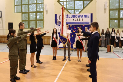 uczniowie klas pierwszych szkoły średniej ślubują na sztandar szkoły. przy sztandarze znajduje się ośmiu uczniów w tym dwóch z klasy o profilu mundurowym. całość odbywa się na hali sportowej.